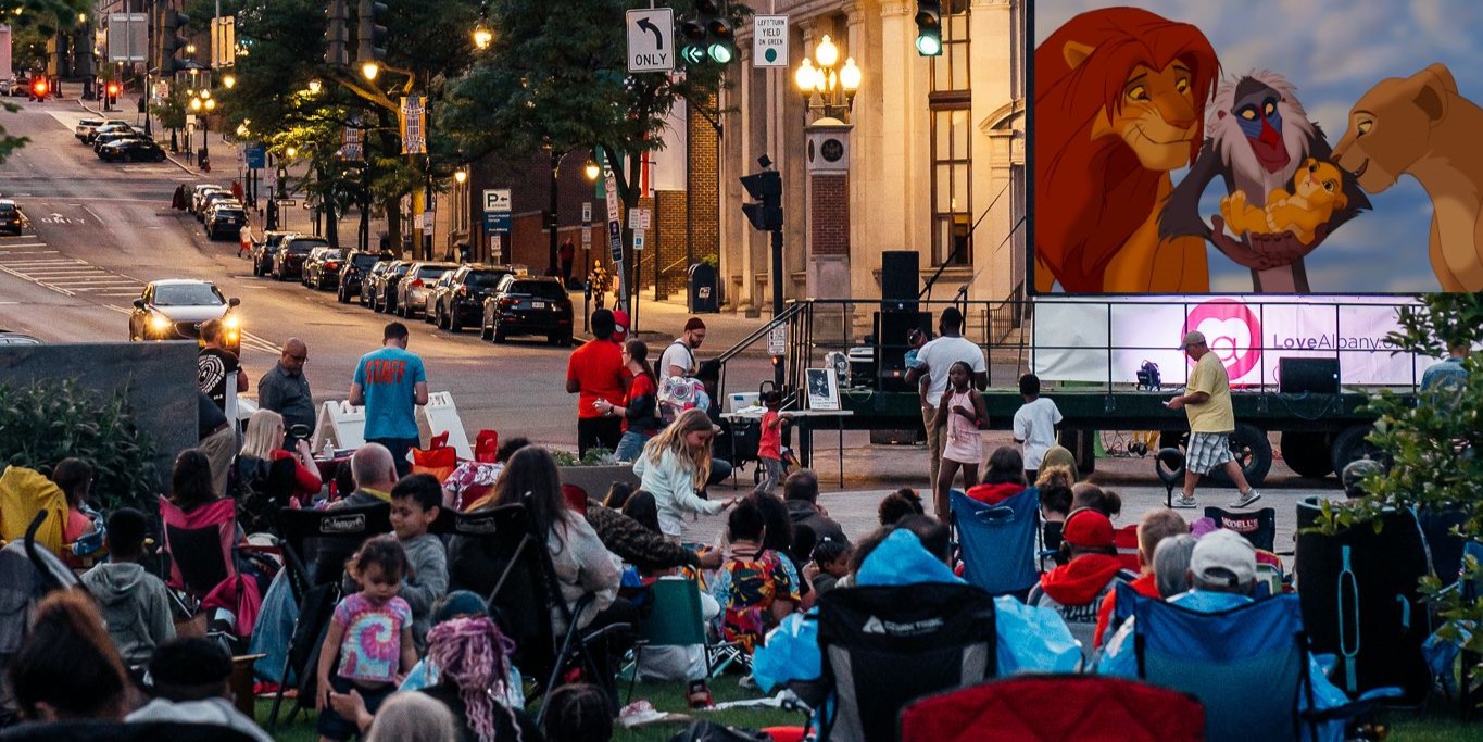 A crowd of people sitting down in lawn chairs and picnic blankets to watch The Lion King (1994) on a large movie screen.