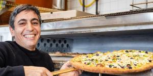 Man, at left, faces camera and smiles as he removes pizza from pizza oven