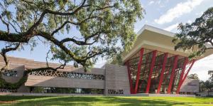 Dynamic image of a striking architectural building with angled red columns holding up a pergola in the front, patterned glass lining the facade. 