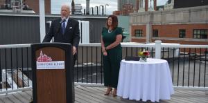 Two people on a rooftop, one standing behind a podium addressing a crowd out of frame, the other standing behind next to a fancy white table