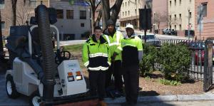 Three employees in reflective jackets stand in front of a park, facing the camera with a smile. A street sweeper on their left. 