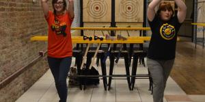 Two people with axes held above their head line up to throw at their targets