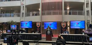 Press event with three screens, a few long tables, and a tv camera 