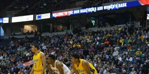 Three basketball players on the court in the middle of a game, the crowd filling the stands behind them. 