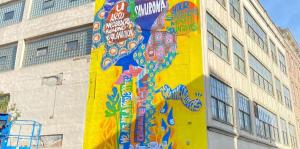 Large mural adorns the side of a building. The illustration is over a bright yellow background and features a mix of portraits and typography. 