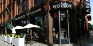 Exterior view of the Hollow restaurant, showing the patio to the left and the front door and main entrance sign to the center. 