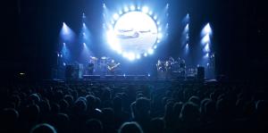 View of a concert stage from the back of the crowd, bright blue lights illuminating the stage and performers. 