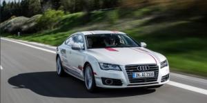 Action shot of a white Audi car on a road, background blurred to indicate speed, the driver raising both hands to show they are not on the steering wheel. 