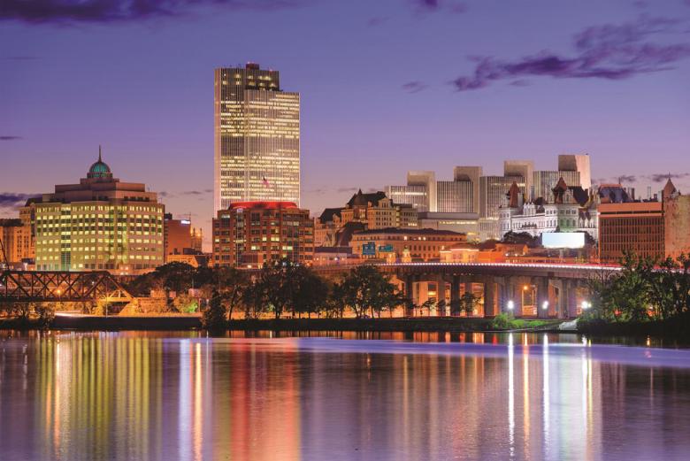 Twilight view of the Albany skyline, building lights reflecting in the still Hudson River in the foreground of the image.