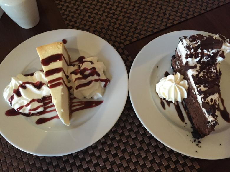 Two plates with decadent slices of cheesecake on each, drizzled with syrups and whipped cream